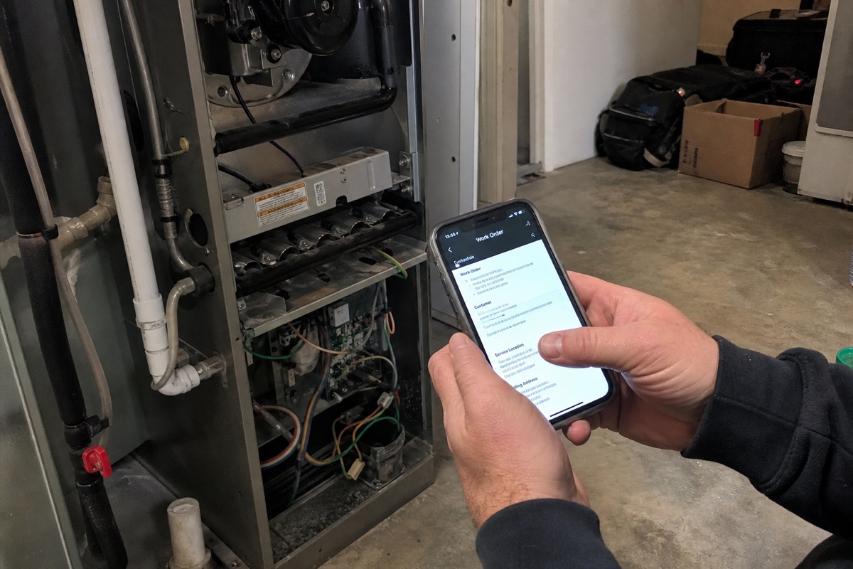 An HVAC tech checking a work order on a phone next to a furnace